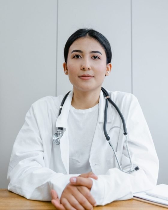 pexels-photo-8376221-8376221 Young female doctor sitting at desk with stethoscope, ready for telehealth consultation.
